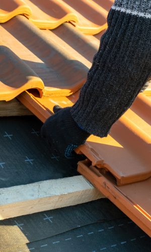 Closeup of worker hands installing yellow ceramic roofing tiles mounted on wooden boards covering residential building roof under construction.